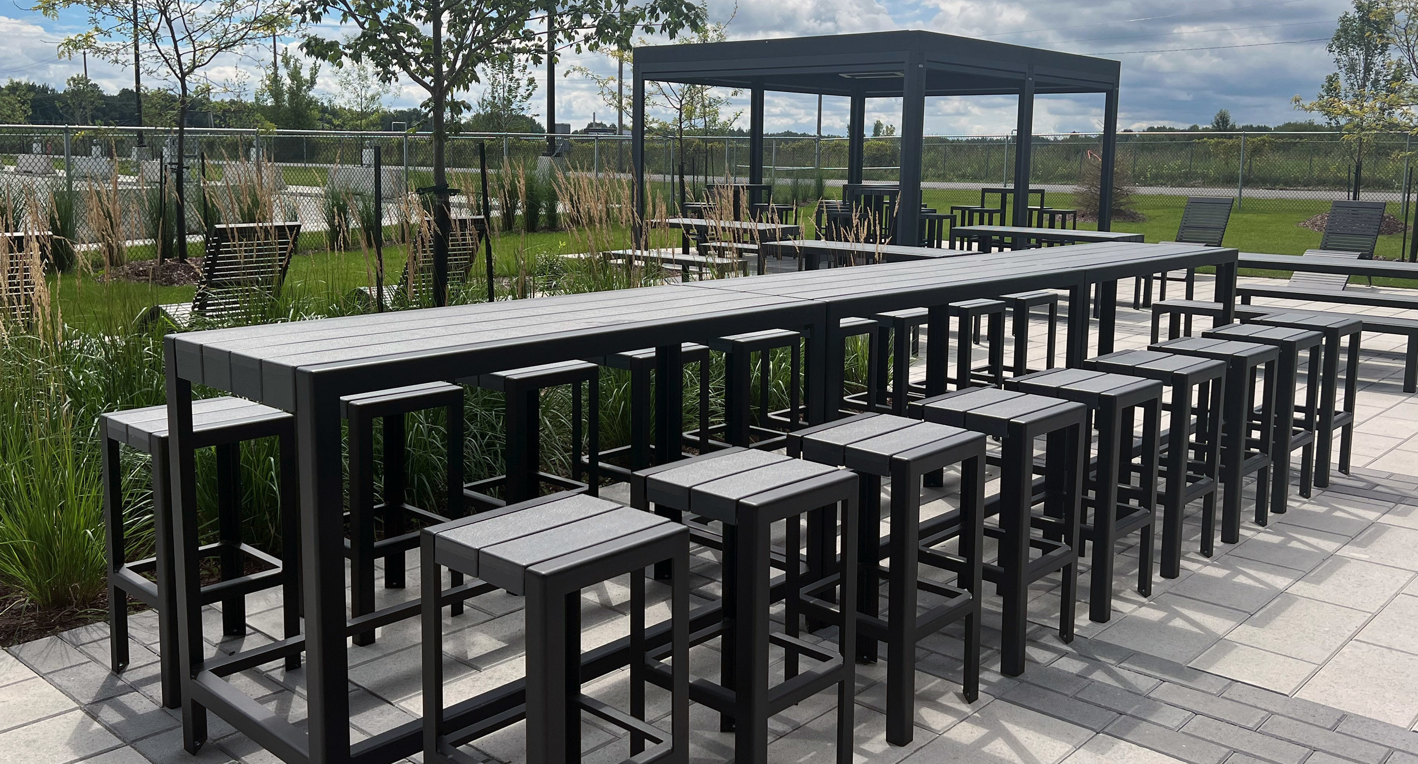 maglin_mtb-1050-bar-height-tables-1 Outdoor patio area with rows of black tables and stools on a paved surface, surrounded by greenery and a fenced boundary under a partly cloudy sky.