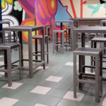 A cafeteria with high square tables and stools on a tiled floor, featuring a colorful mural on the back wall.