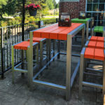 Outdoor patio with modern tables and stools featuring orange and green tops, metal frames, and a brick building in the background. Lush greenery and a flowerpot are visible.