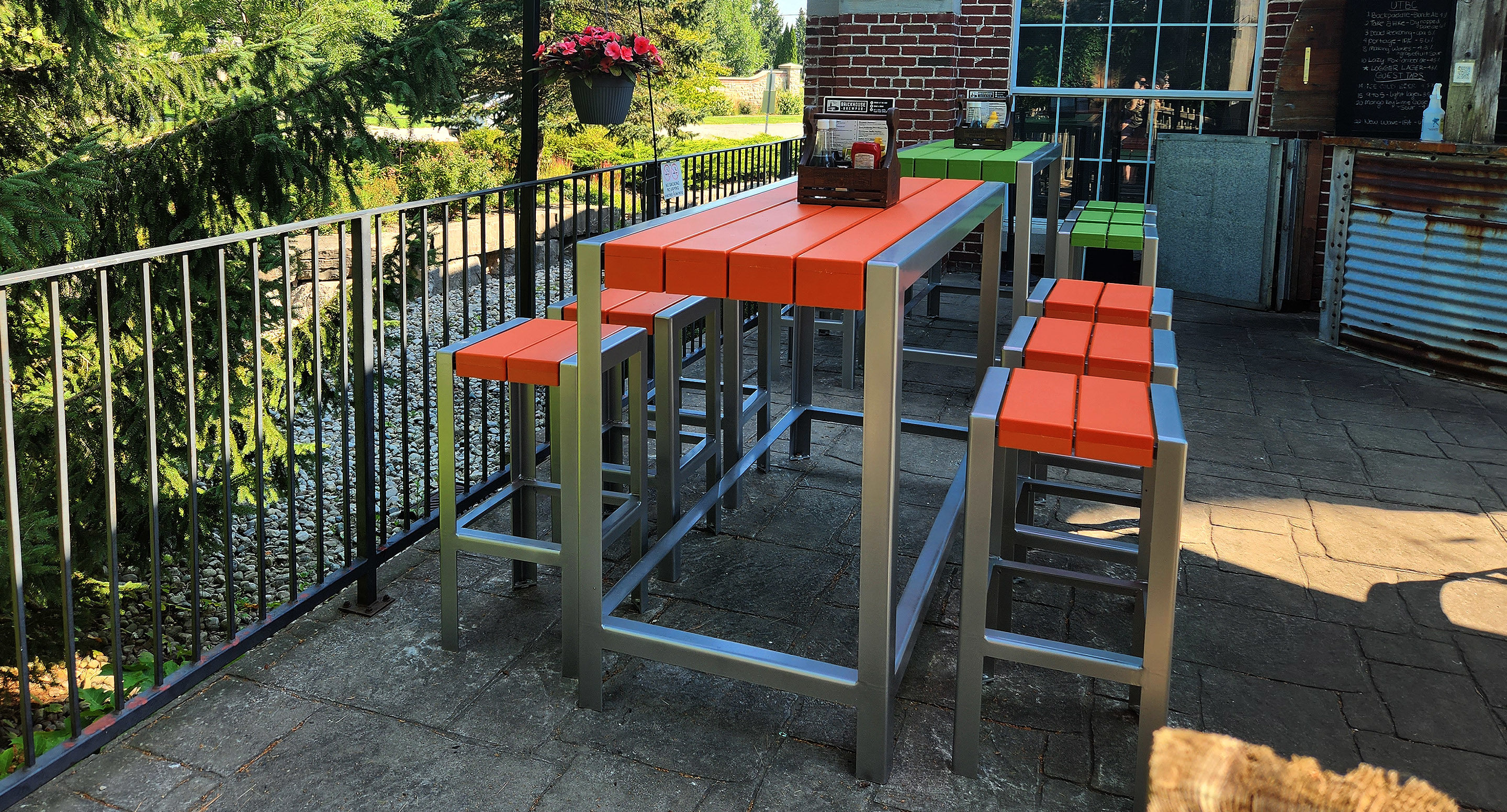 maglin_mtb-1050-bar-height-tables-3 Outdoor patio with modern tables and stools featuring orange and green tops, metal frames, and a brick building in the background. Lush greenery and a flowerpot are visible.