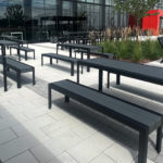 Outdoor patio area with black metal tables, benches, and lounge chairs on light stone pavers next to a modern glass building and landscaped plants.