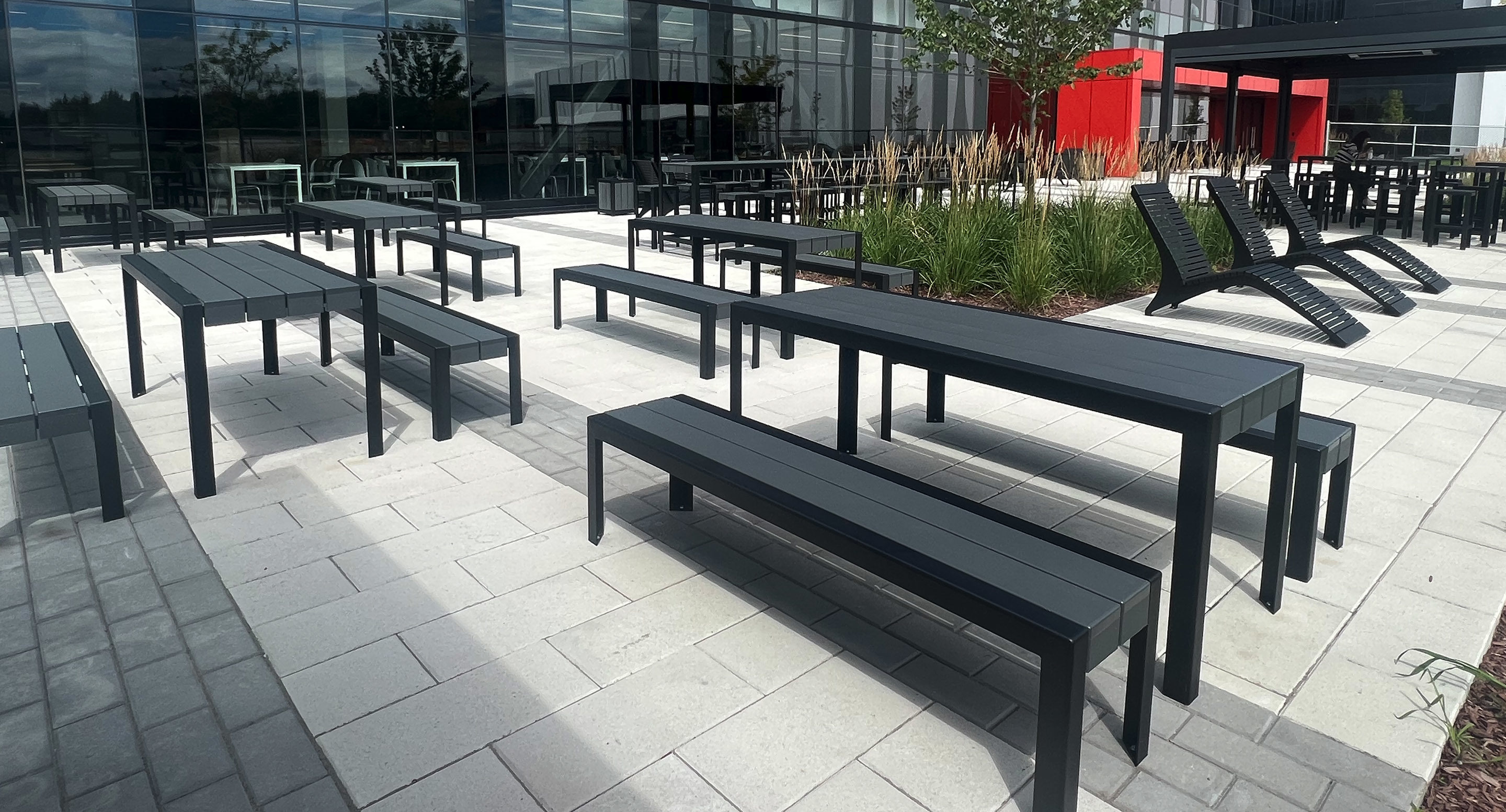 Outdoor patio area with black metal tables, benches, and lounge chairs on light stone pavers next to a modern glass building and landscaped plants.