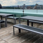 Rooftop patio with metal picnic tables and benches on wooden decking, overlooking a city skyline under a cloudy sky.