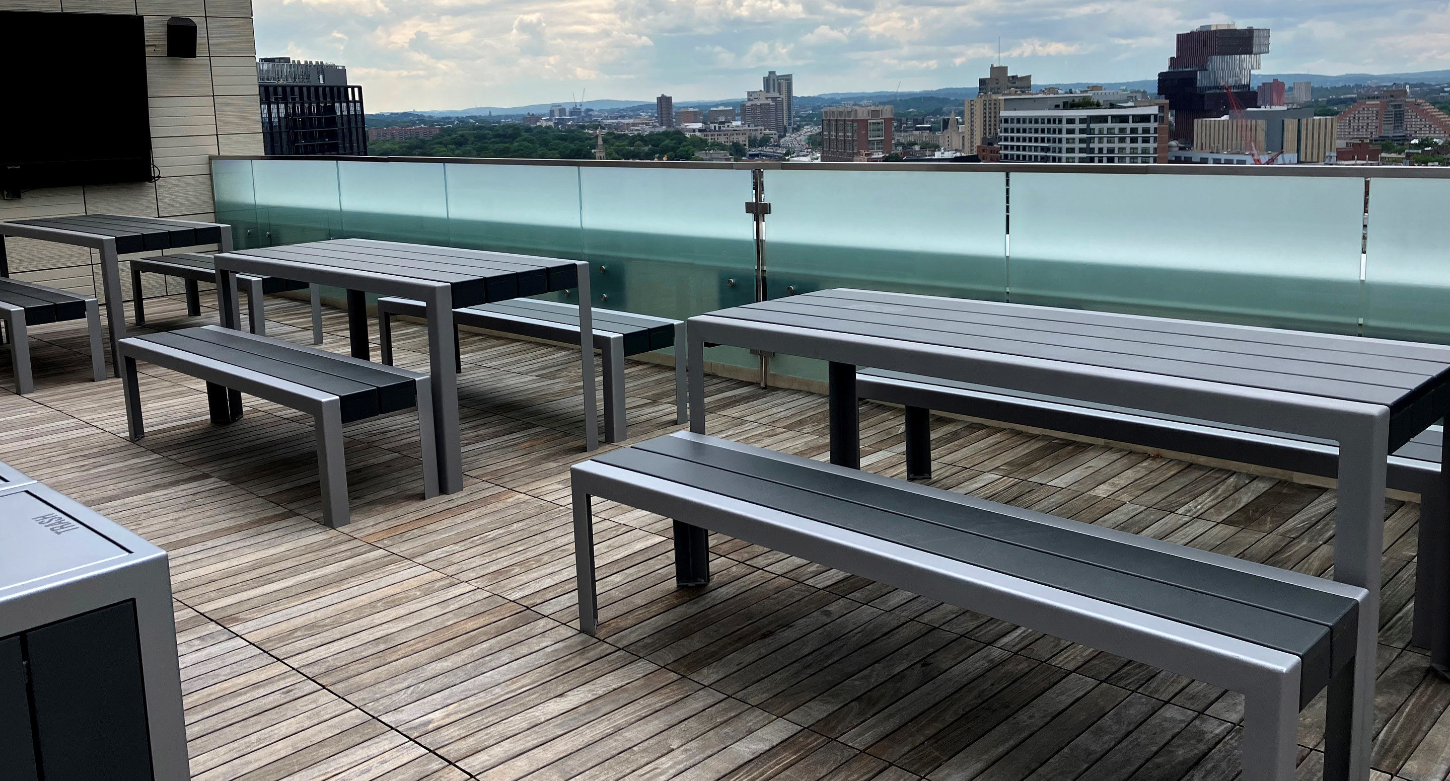 maglin_mtb-1050-standard-tables-2 Rooftop patio with metal picnic tables and benches on wooden decking, overlooking a city skyline under a cloudy sky.