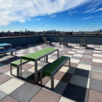 Rooftop patio with green and blue picnic tables, chairs, and city skyline view under a partly cloudy sky.