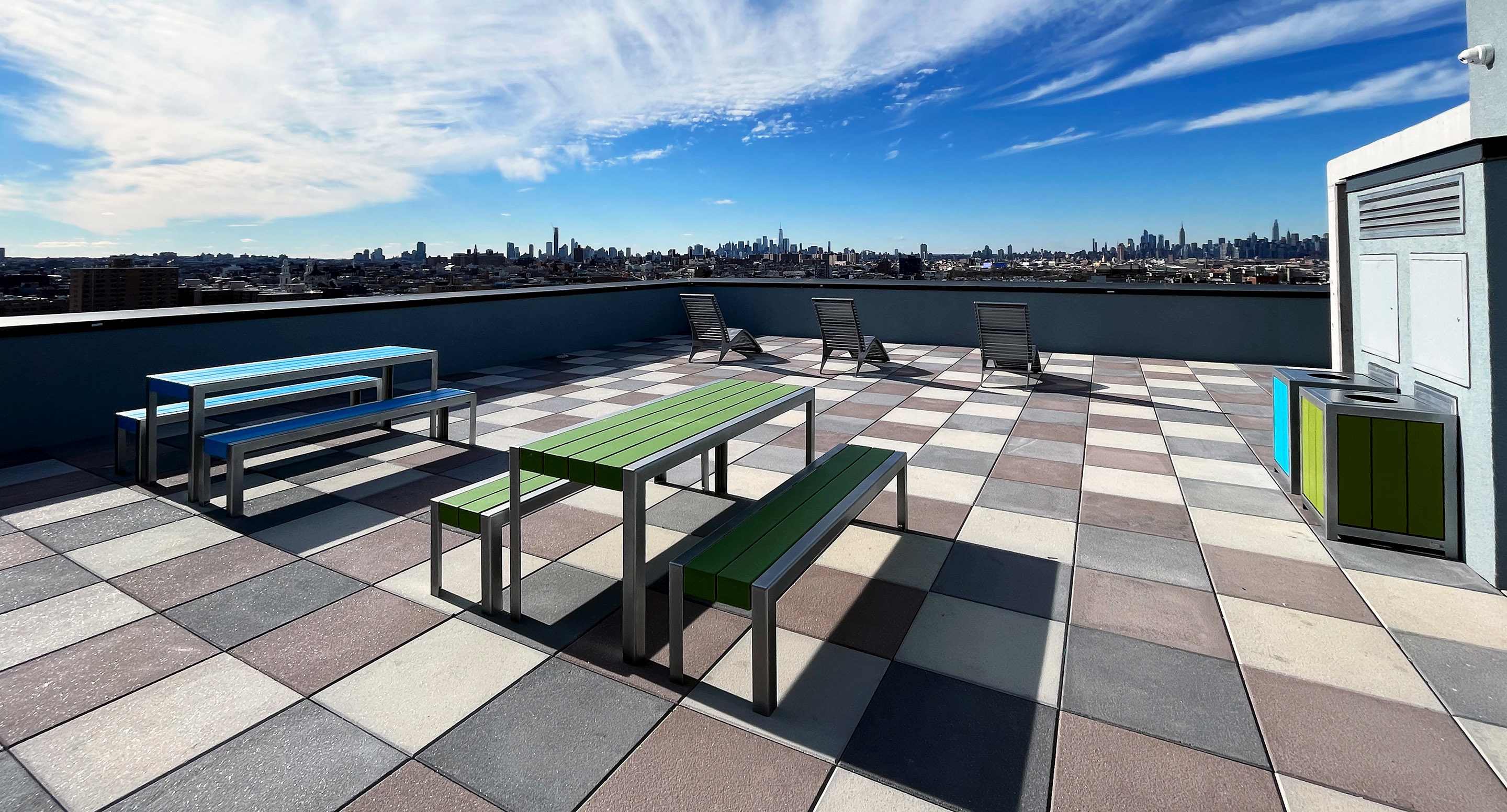 maglin_mtb-1050-standard-tables-3 Rooftop patio with green and blue picnic tables, chairs, and city skyline view under a partly cloudy sky.