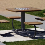 Une table de pique-nique en bois avec quatre sièges attachés et un motif d'échiquier sur le plateau, située sur une aire en béton entourée d'herbe.