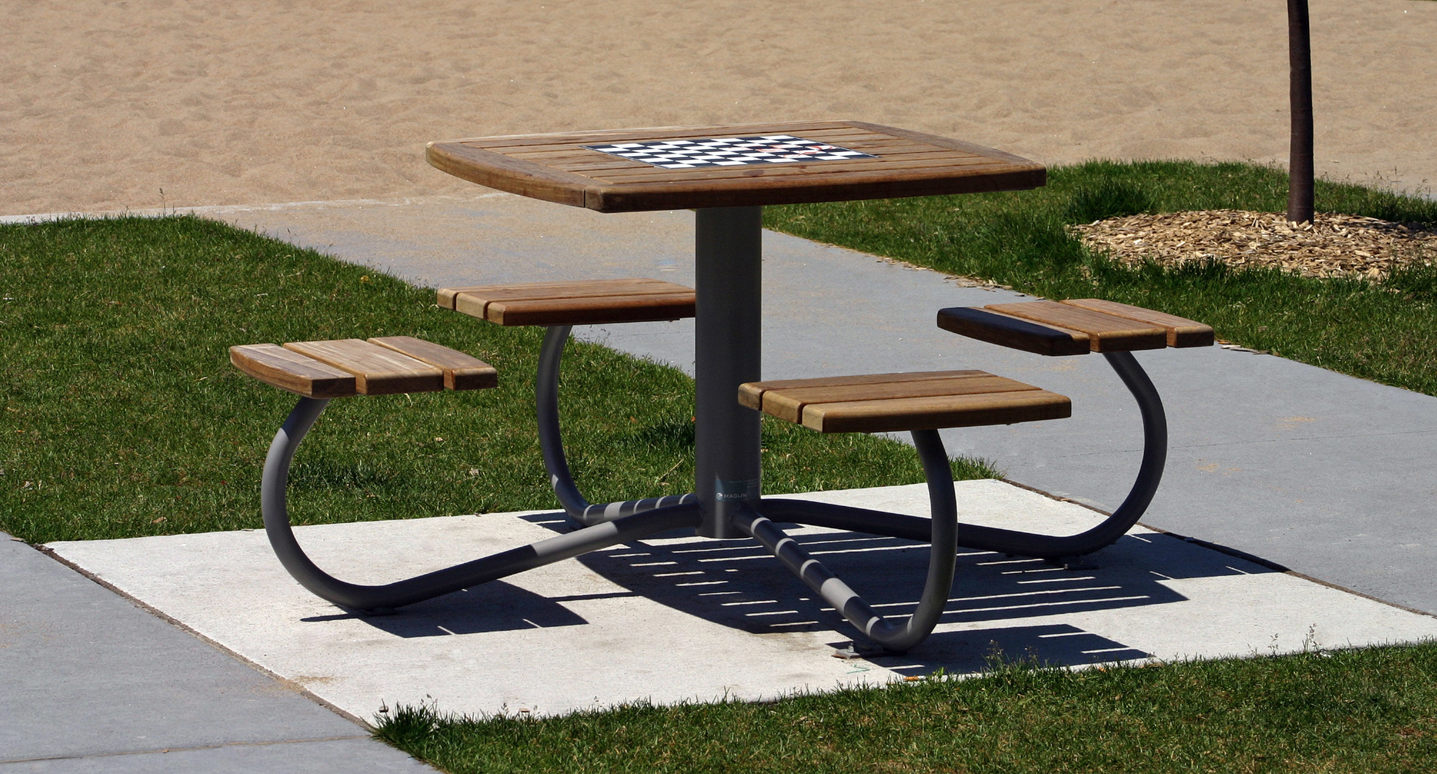 maglin_mtb-1100-cluster-seating-1 Une table de pique-nique en bois avec quatre sièges attachés et un motif d'échiquier sur le plateau, située sur une aire en béton entourée d'herbe.