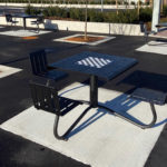 Table de pique-nique en plein air avec des bancs attenants et un échiquier intégré, située dans une zone pavée pratiquement vide avec d'autres tables similaires et quelques arbres nouvellement plantés.