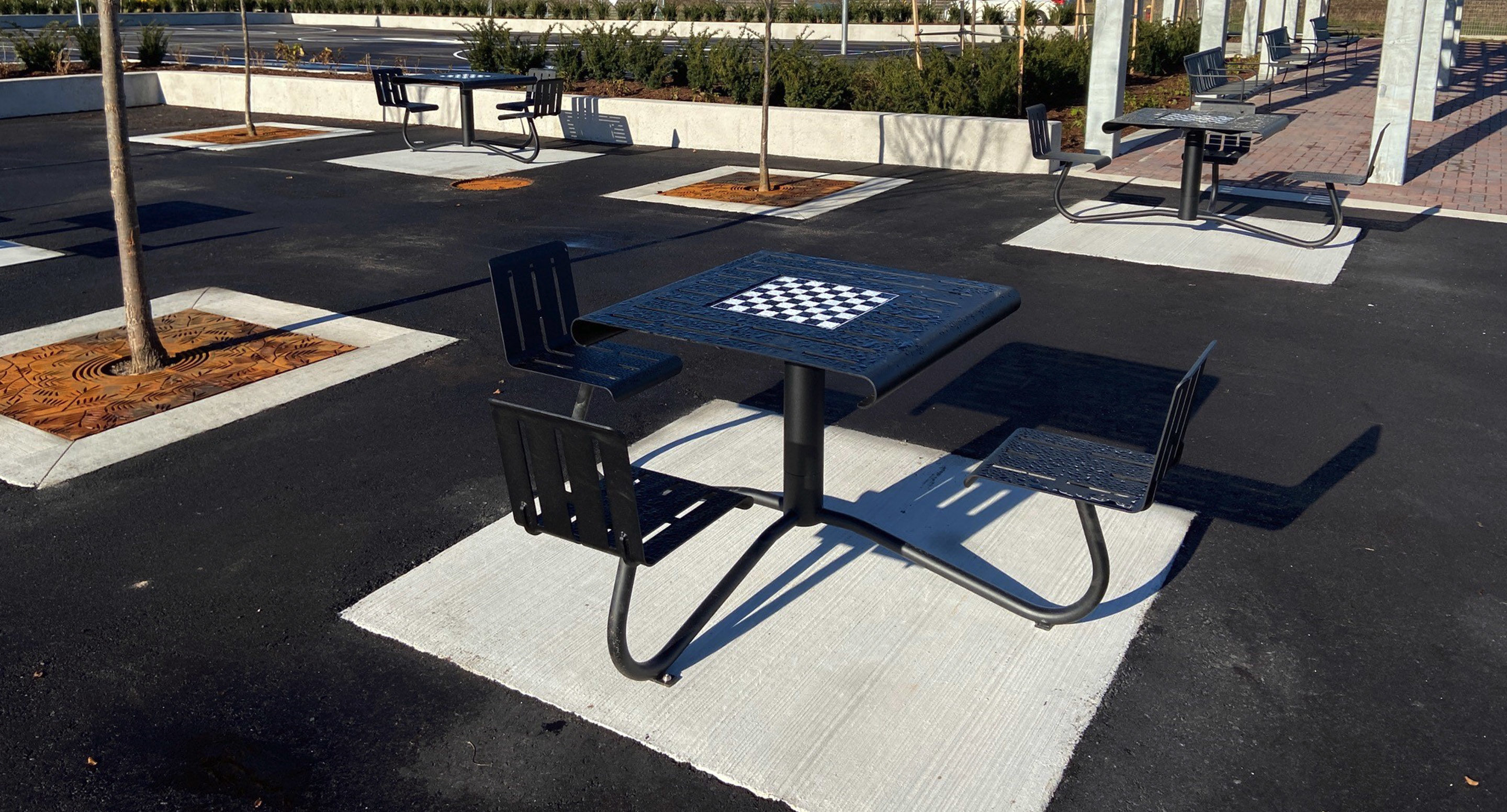 maglin_mtb-1100-cluster-seating-2 Table de pique-nique en plein air avec des bancs attenants et un échiquier intégré, située dans une zone pavée pratiquement vide avec d'autres tables similaires et quelques arbres nouvellement plantés.