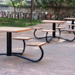 Plusieurs tables de pique-nique en bois avec leurs bancs sont disposées sur une aire pavée à l'extérieur, avec de l'herbe, des arbres et une rangée de bicyclettes garées à l'arrière-plan.