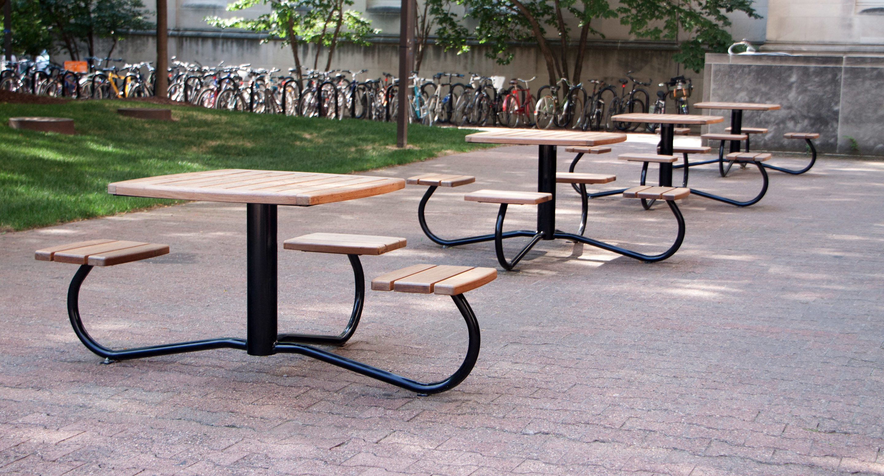 maglin_mtb-1100-cluster-seating-4 Plusieurs tables de pique-nique en bois avec leurs bancs sont disposées sur une aire pavée à l'extérieur, avec de l'herbe, des arbres et une rangée de bicyclettes garées à l'arrière-plan.