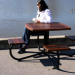 Une personne assise seule à une table de pique-nique en bois près d'un plan d'eau, face à des canards qui nagent en arrière-plan.