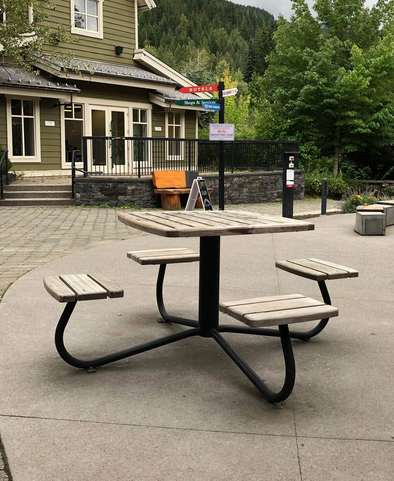 La Table et sièges groupés 1100, une table de pique-nique carrée en bois avec quatre bancs attachés, est située sur une zone pavée près d'un bâtiment et entourée d'arbres.