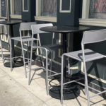 Three tall tables with pairs of gray chairs are arranged outside a building with large windows and dark green walls, casting shadows on the sidewalk.