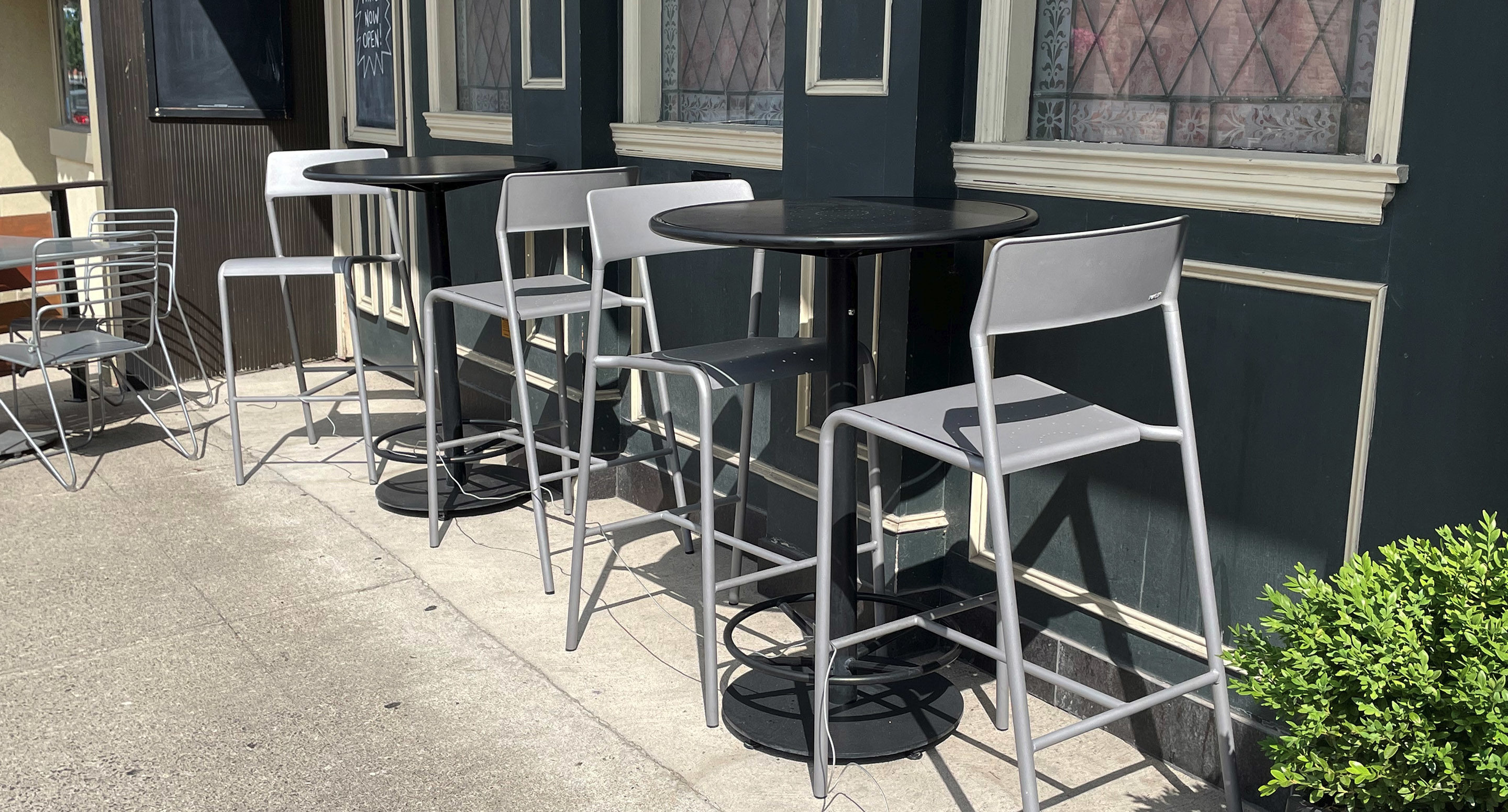 Three tall tables with pairs of gray chairs are arranged outside a building with large windows and dark green walls, casting shadows on the sidewalk.
