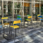 Outdoor seating area with several square tables and colorful chairs in blue, yellow, and green, set on a tiled floor near large windows and potted plants.