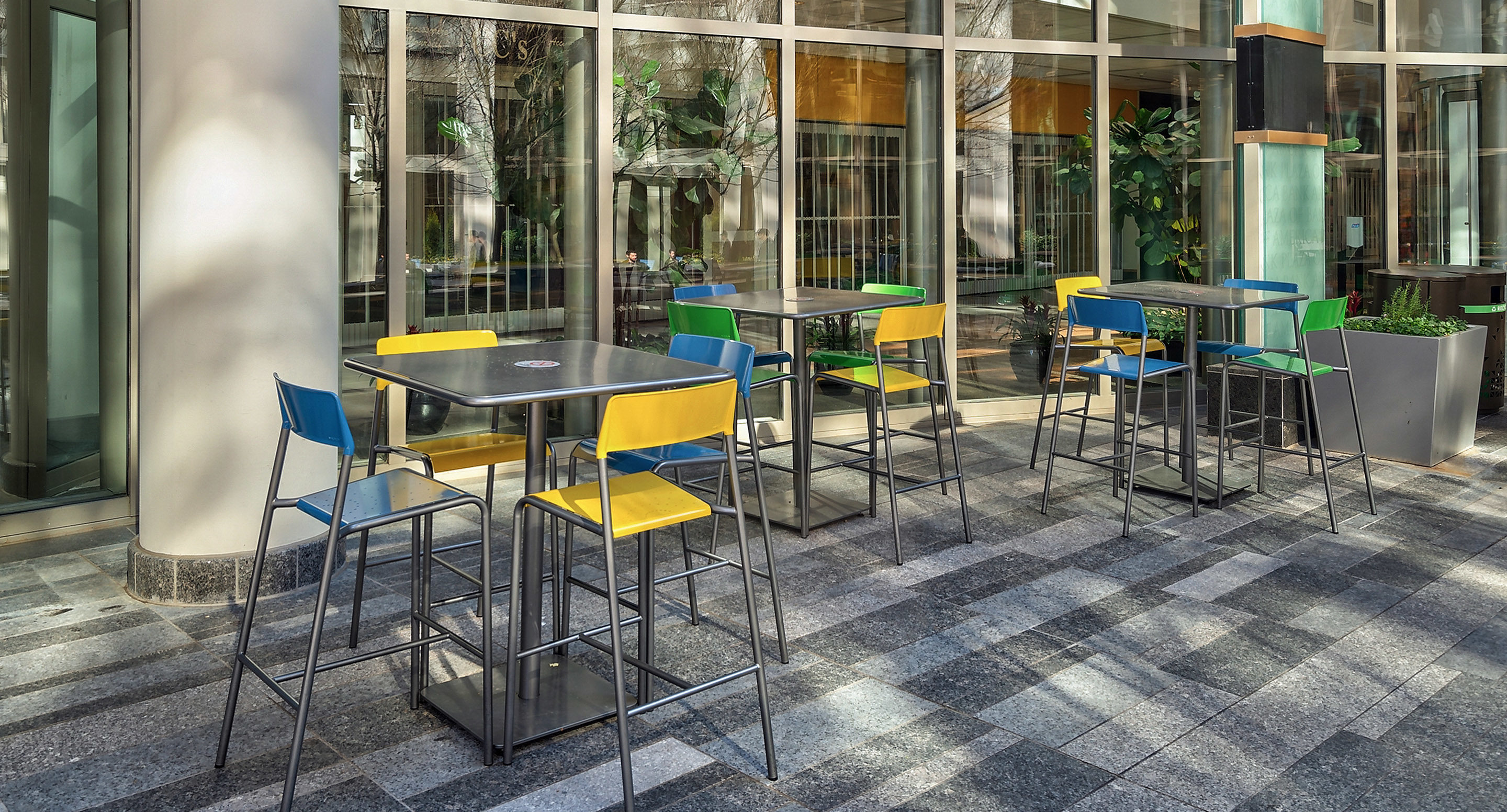 Outdoor seating area with several square tables and colorful chairs in blue, yellow, and green, set on a tiled floor near large windows and potted plants.
