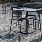 A metal table with four high chairs sits on a gray brick patio in an outdoor seating area with more tables and chairs in the background.