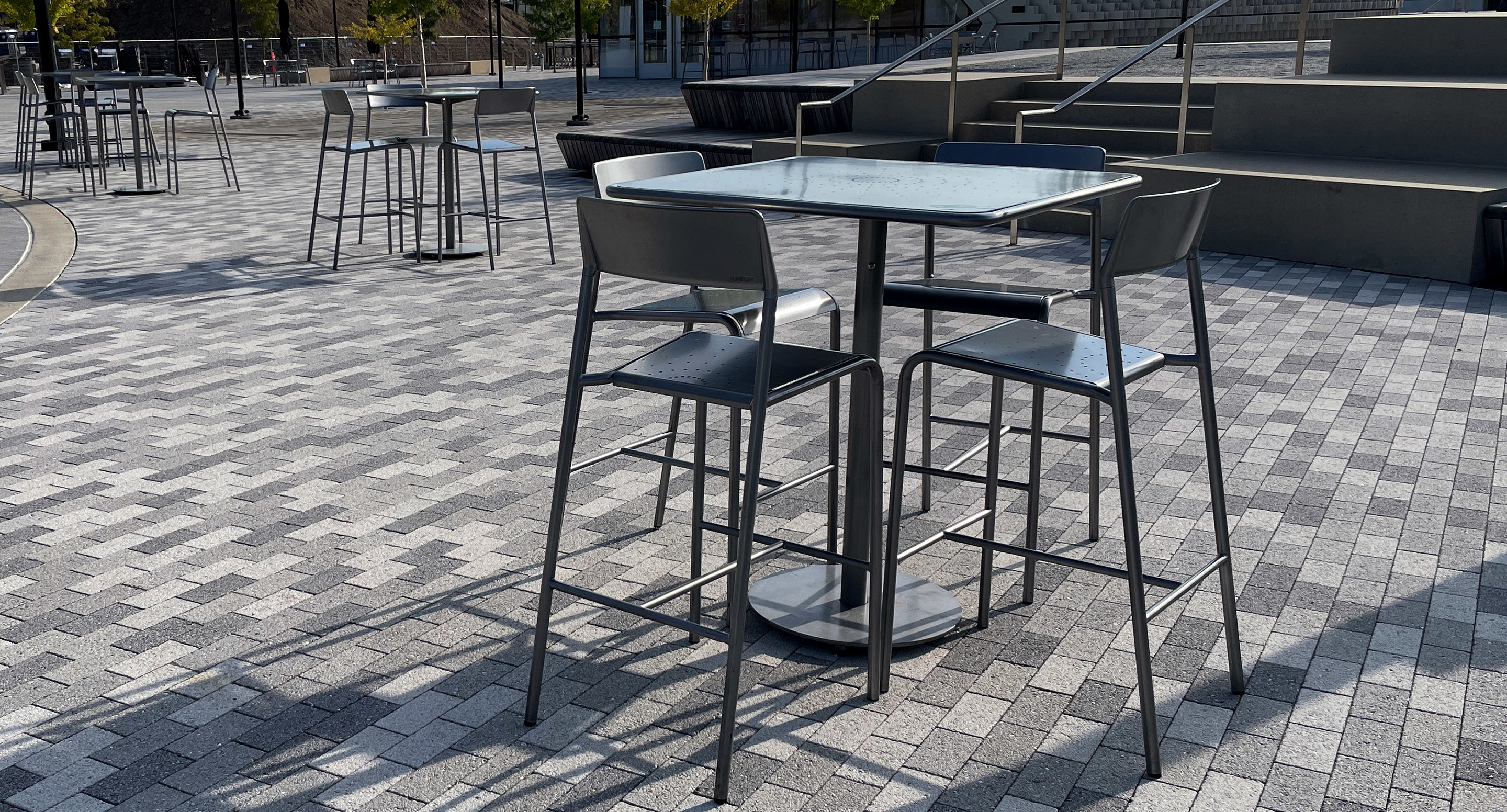 A metal table with four high chairs sits on a gray brick patio in an outdoor seating area with more tables and chairs in the background.