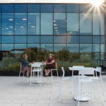 Two people sit at a white table on a rooftop patio with modern glass building facade behind them and sun shining through upper corner.