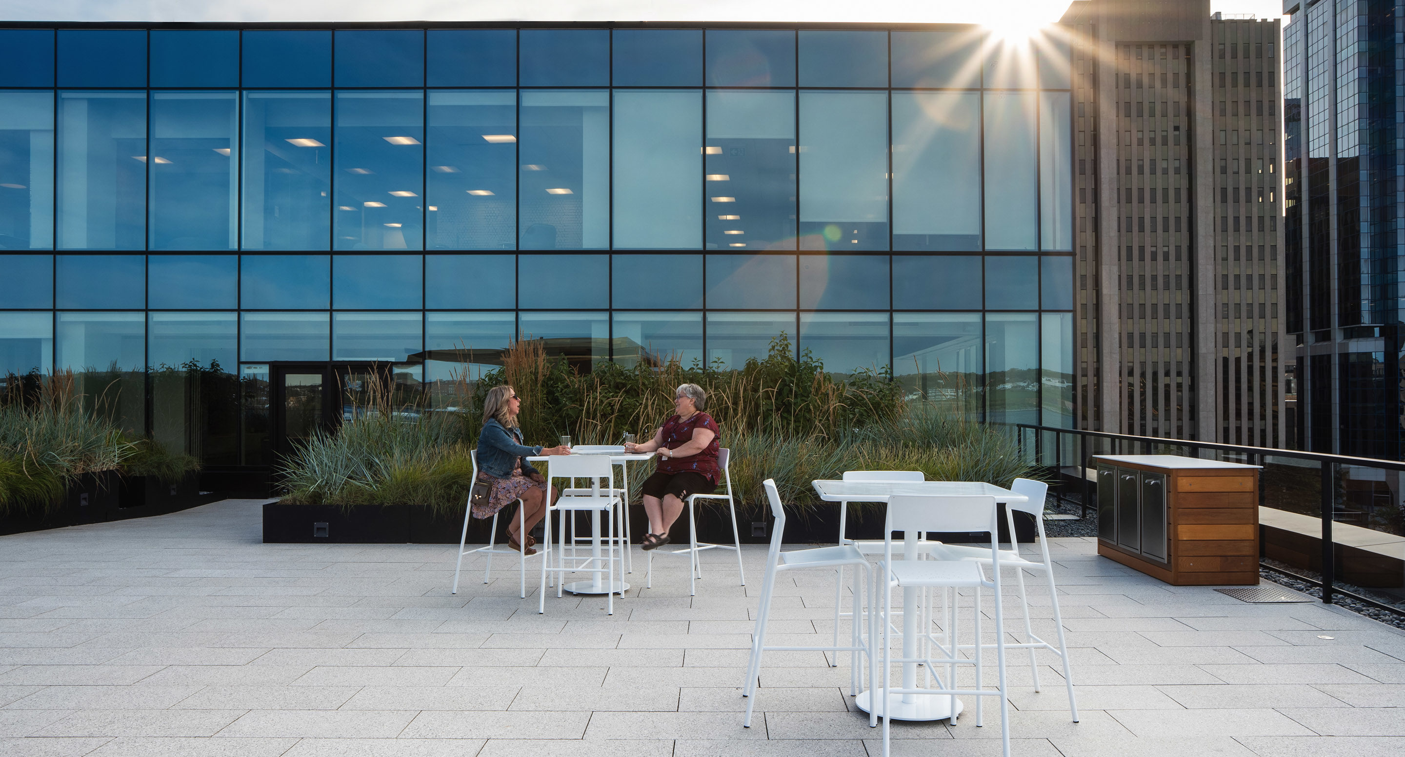 Two people sit at a white table on a rooftop patio with modern glass building facade behind them and sun shining through upper corner.