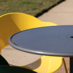A round metal outdoor table with a yellow and a green chair on a sunny day, set next to a sidewalk and grass.