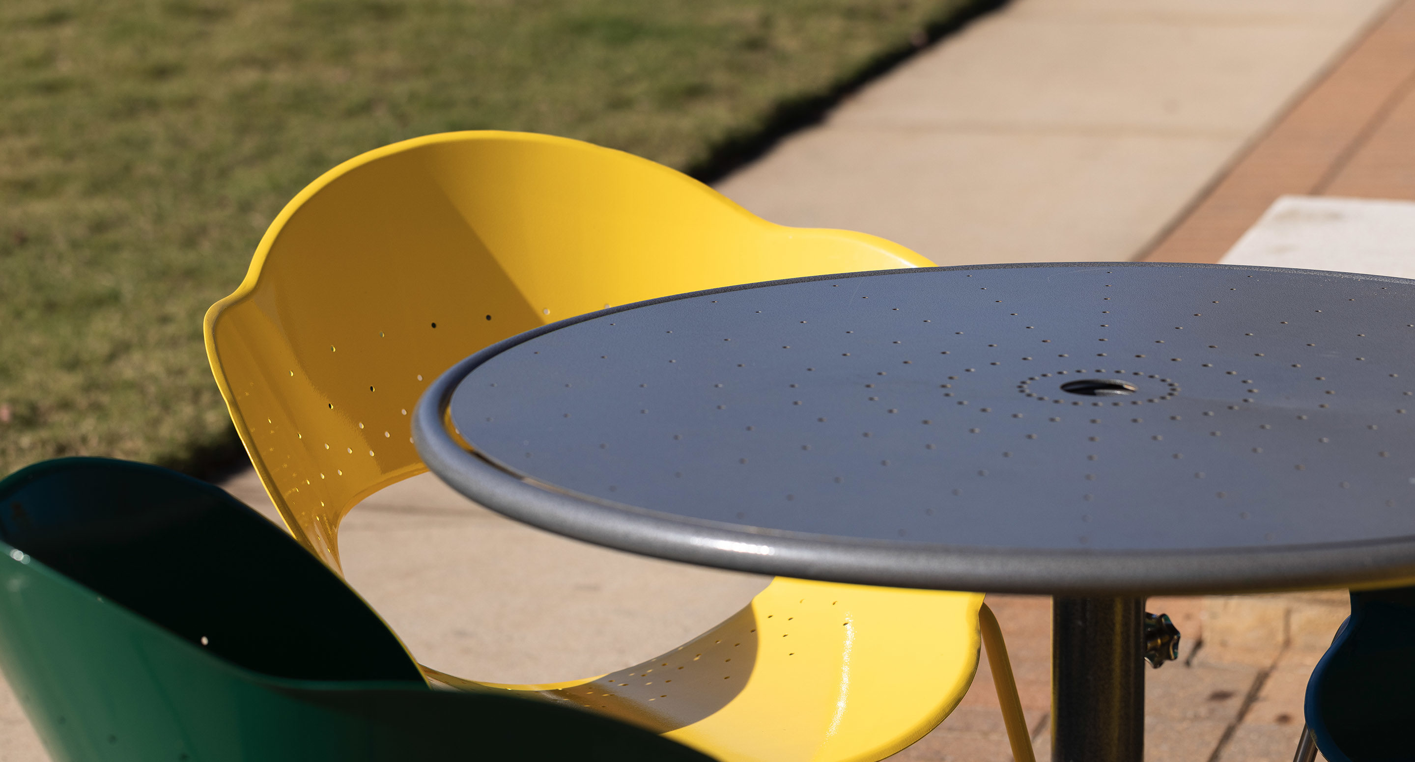 maglin_mtb-1700-foro-dining-tables-1 A round metal outdoor table with a yellow and a green chair on a sunny day, set next to a sidewalk and grass.