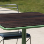 A square wooden table with a green frame is accompanied by two metal chairs, situated outdoors on a concrete surface with grass in the background.