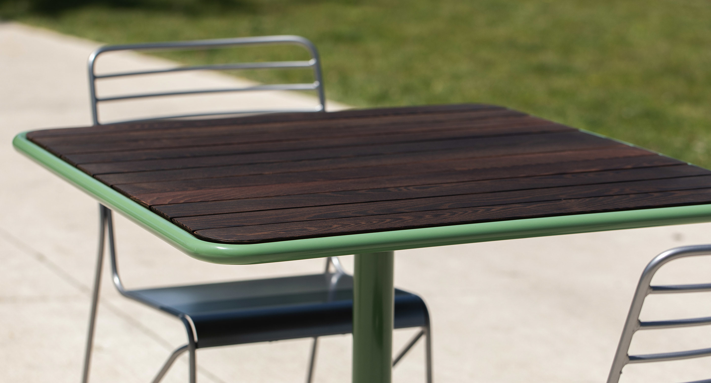 maglin_mtb-1700-foro-dining-tables-2 A square wooden table with a green frame is accompanied by two metal chairs, situated outdoors on a concrete surface with grass in the background.