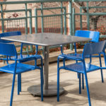 A square metal table with four blue chairs is arranged on a concrete patio next to a green metal railing.