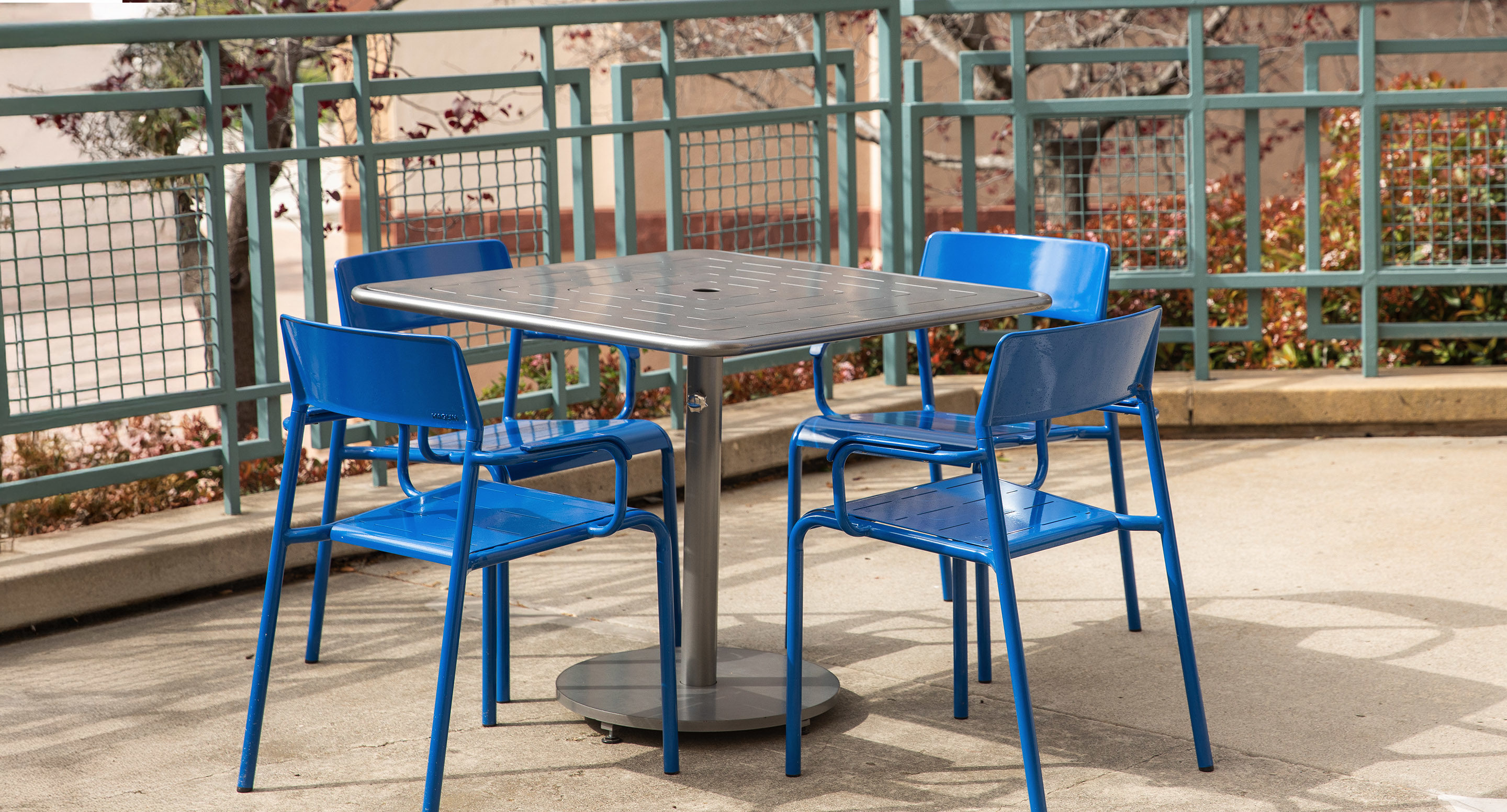 maglin_mtb-1700-foro-dining-tables-3 A square metal table with four blue chairs is arranged on a concrete patio next to a green metal railing.