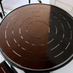 A round, dark-colored metal table with concentric cut-out lines is shown on a light-colored surface, accompanied by two matching chairs.