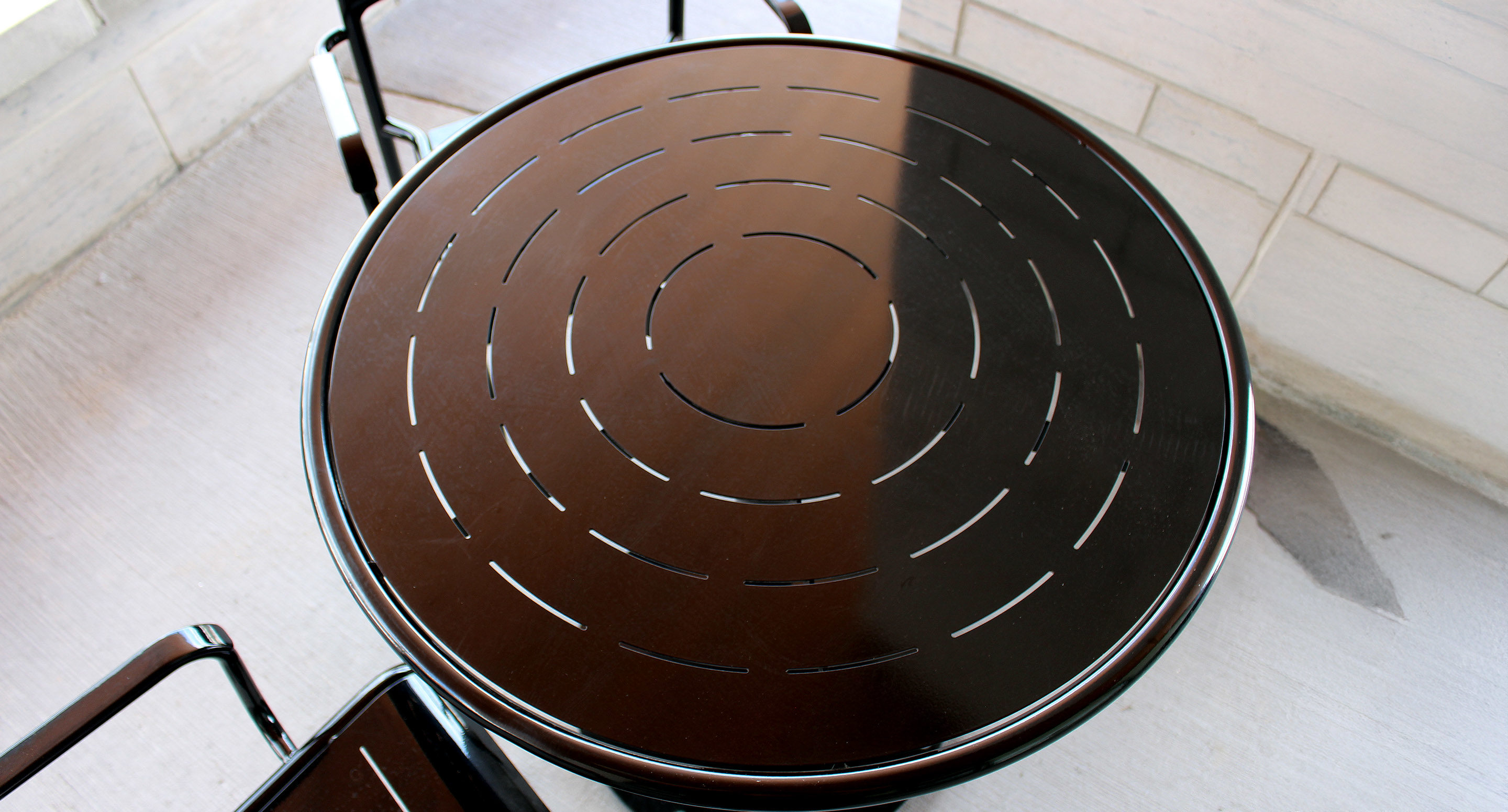 maglin_mtb-1700-foro-dining-tables-4 A round, dark-colored metal table with concentric cut-out lines is shown on a light-colored surface, accompanied by two matching chairs.