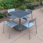 A metal table with four matching chairs is placed on a paved outdoor area next to some greenery, with additional tables and chairs in the background.