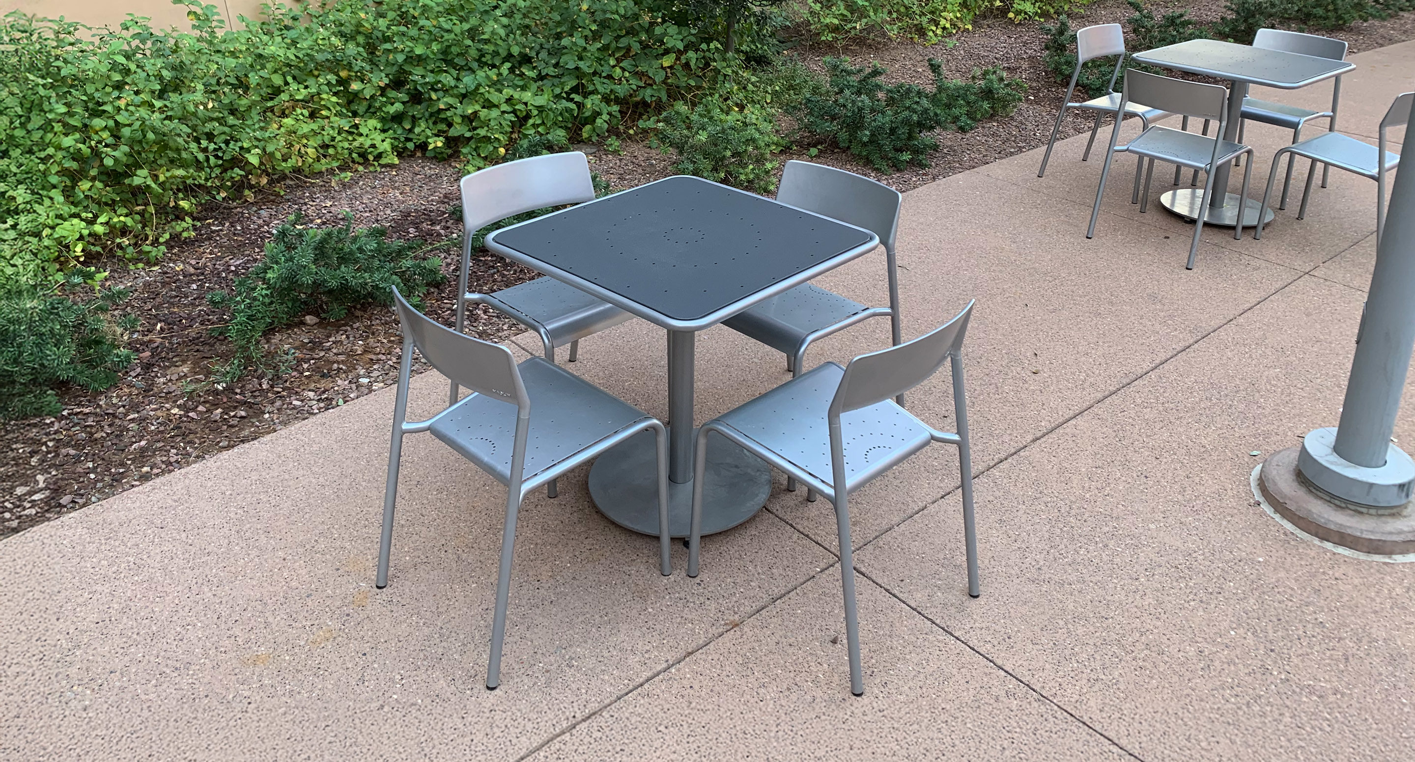 maglin_mtb-1700-foro-dining-tables-5 A metal table with four matching chairs is placed on a paved outdoor area next to some greenery, with additional tables and chairs in the background.