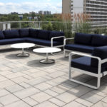 Modern outdoor patio with two navy cushioned sofas, two round white tables, and tall grasses, set on a tiled rooftop with city buildings and greenery in the background.