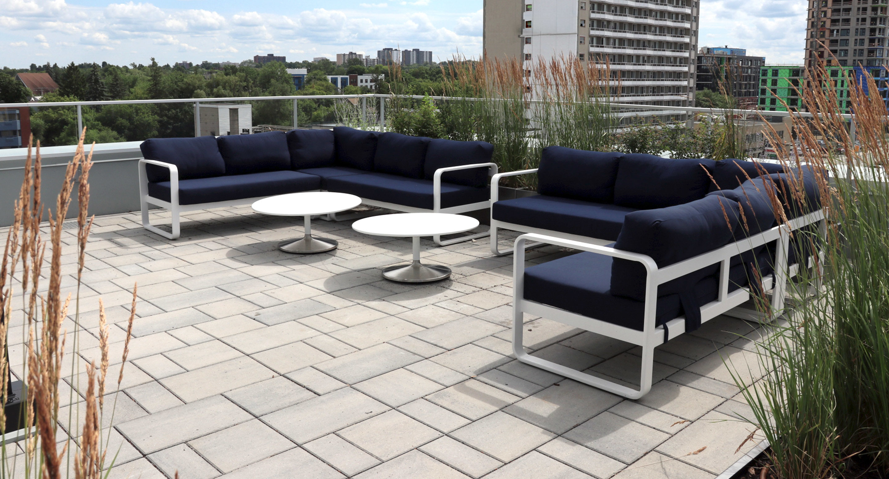 Modern outdoor patio with two navy cushioned sofas, two round white tables, and tall grasses, set on a tiled rooftop with city buildings and greenery in the background.