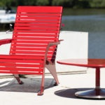 A red metal lounge chair and a matching round table sit on a concrete patio beside a body of water, with a boat and trees visible in the background.
