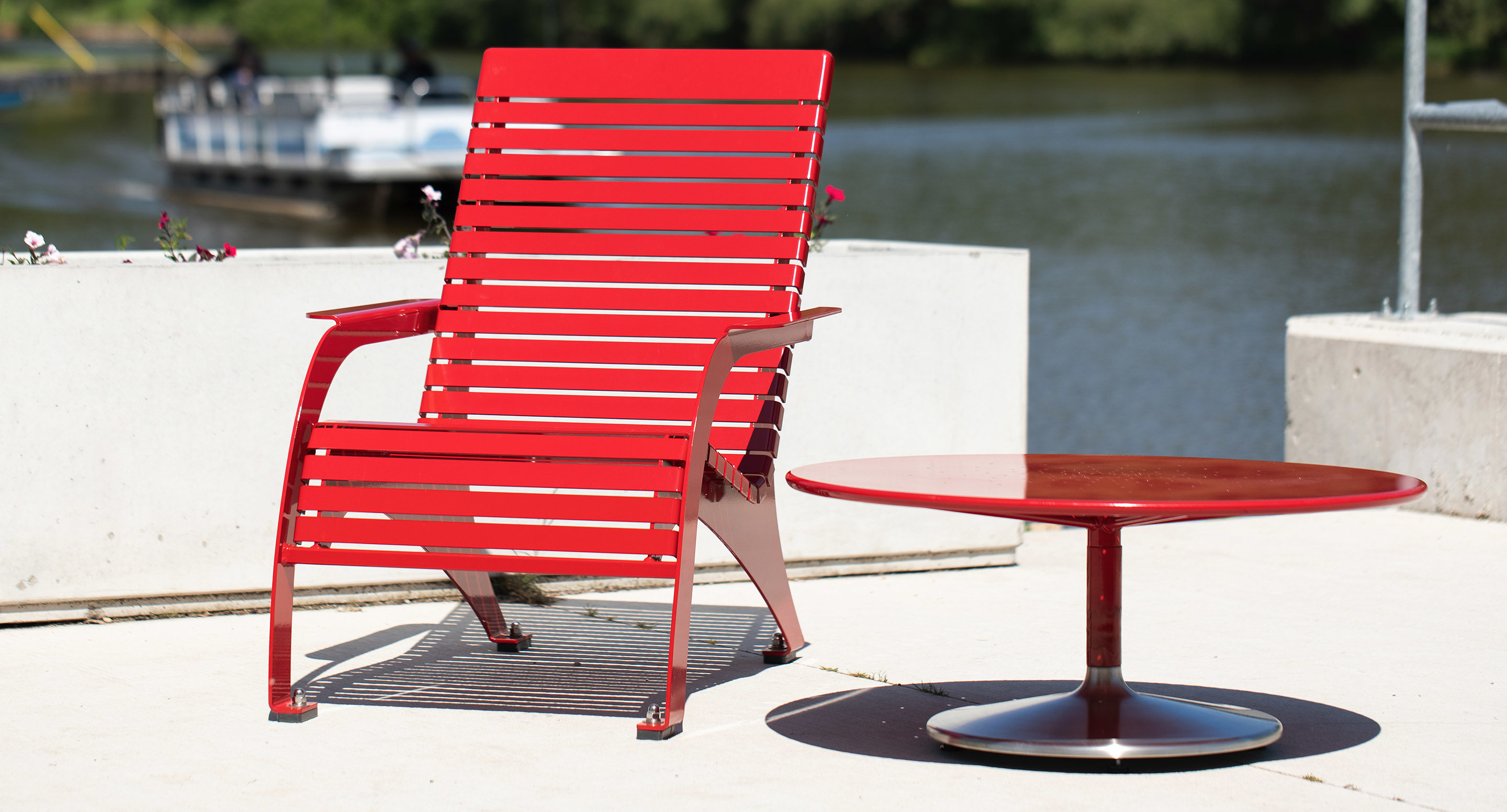 A red metal lounge chair and a matching round table sit on a concrete patio beside a body of water, with a boat and trees visible in the background.