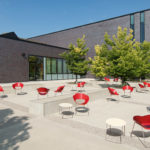 Outdoor patio area with scattered red chairs and small round tables, surrounded by trees and adjacent to a modern brick building under a clear sky.