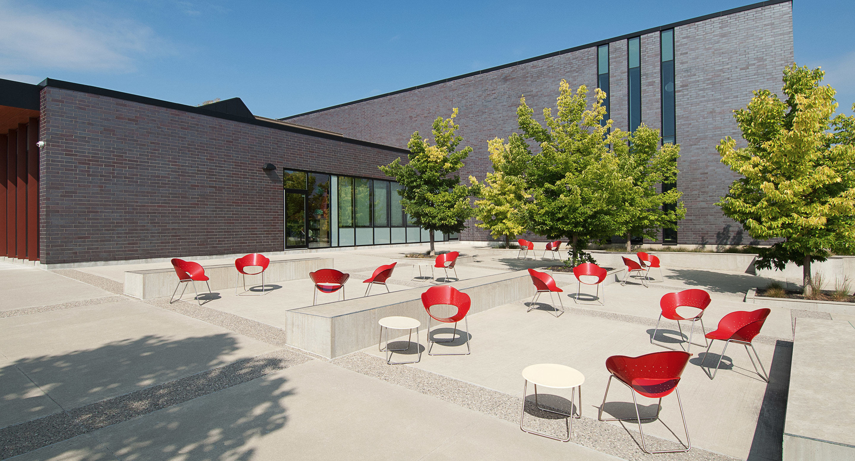 Outdoor patio area with scattered red chairs and small round tables, surrounded by trees and adjacent to a modern brick building under a clear sky.