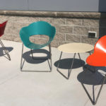 Three colorful chairs (red, teal, and orange) and a round beige table are arranged on a concrete patio in front of a stone wall and blue panel.