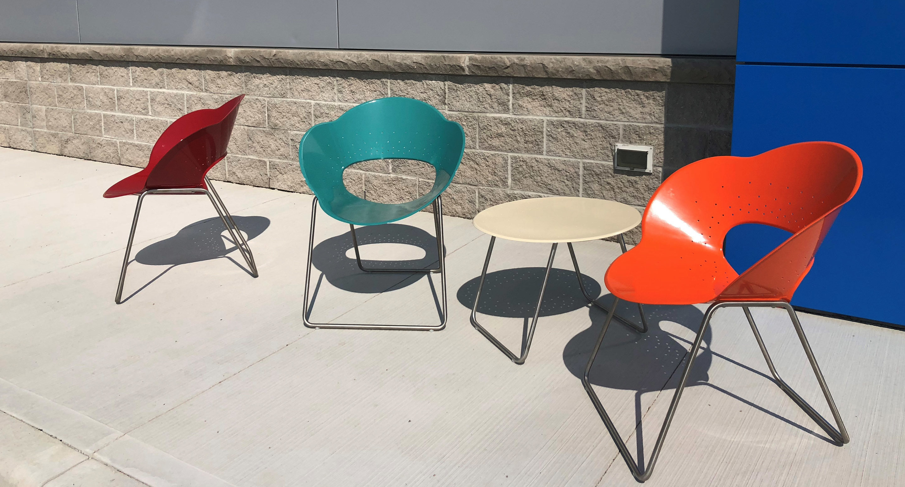 Three colorful chairs (red, teal, and orange) and a round beige table are arranged on a concrete patio in front of a stone wall and blue panel.