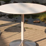 Une table ronde blanche avec une base en métal est assise entre deux chaises orange sur une surface en béton à côté d'un plan d'eau avec des algues vertes et des rochers.