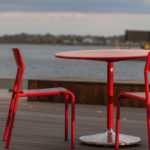 Deux chaises métalliques rouges et une table ronde assortie sont placées sur une terrasse en bois au bord de l'eau, avec des bâtiments et de l'eau visibles à l'arrière-plan.