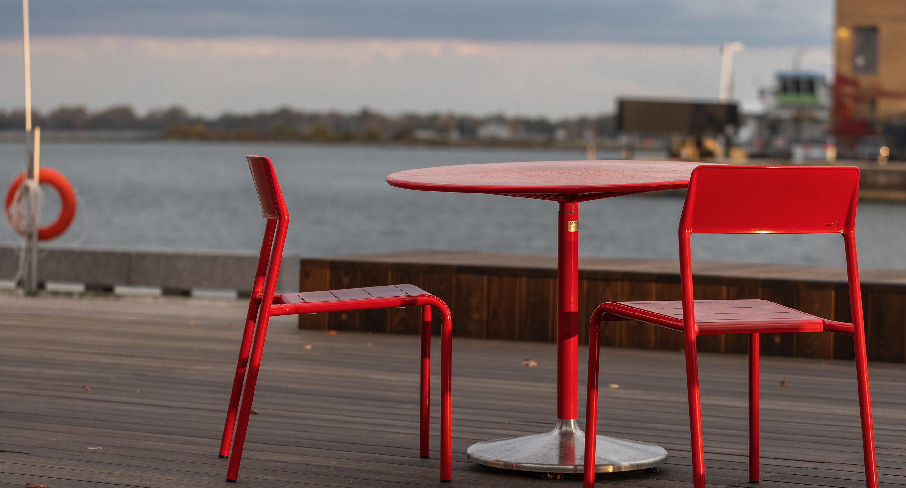 maglin_mtb-1800-battery-tables-3 Deux chaises métalliques rouges et une table ronde assortie sont placées sur une terrasse en bois au bord de l'eau, avec des bâtiments et de l'eau visibles à l'arrière-plan.