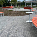 Outdoor park with curved orange benches, round white tables, paved walkways, scattered trees, and people sitting in the background near a water feature.