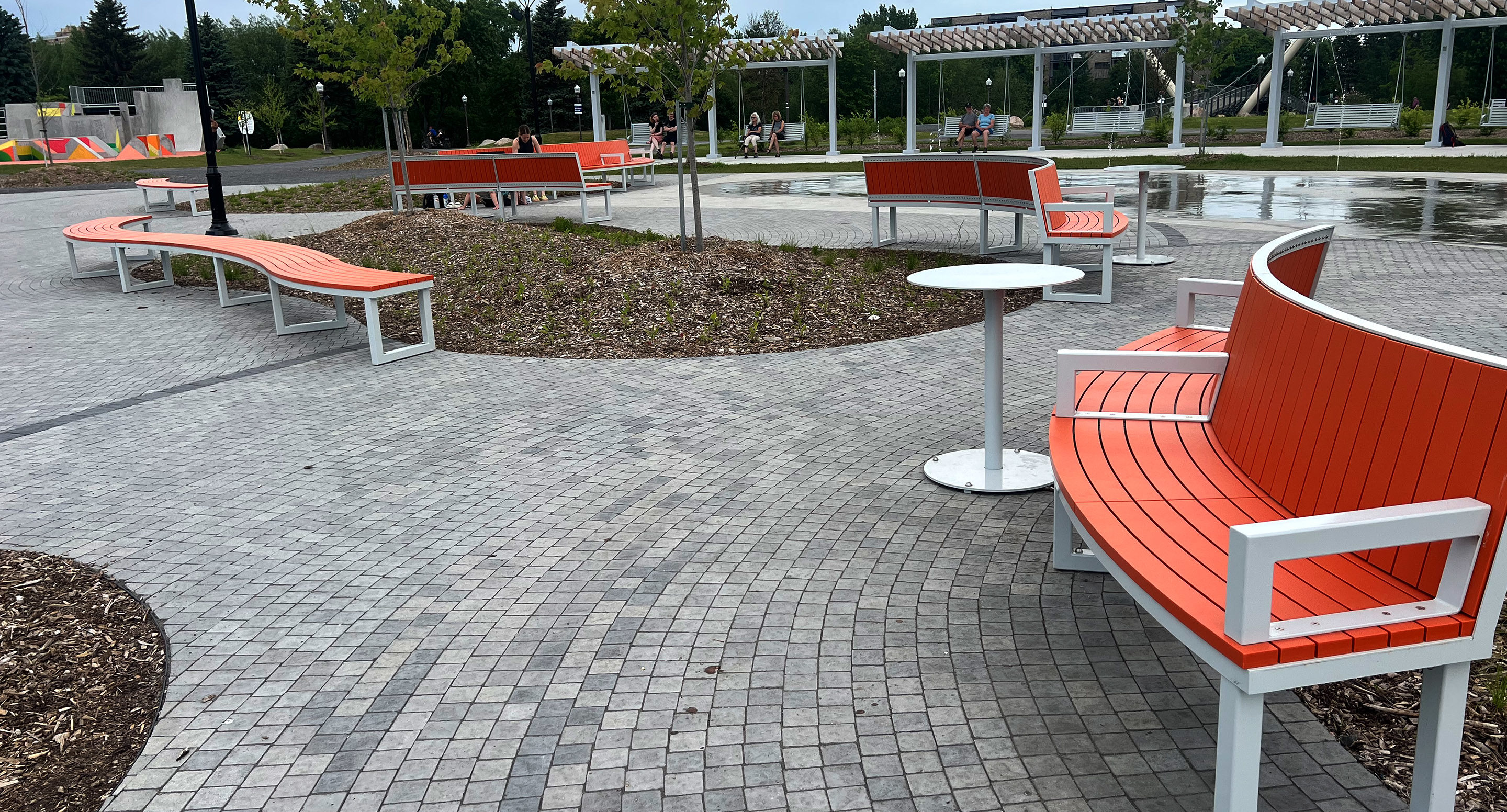 maglin_mtb-1900-ogden-cafe-tables-1 Outdoor park with curved orange benches, round white tables, paved walkways, scattered trees, and people sitting in the background near a water feature.
