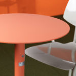 A round, coral-colored table is next to a white metal chair with a contoured seat on a green floor with an orange background.